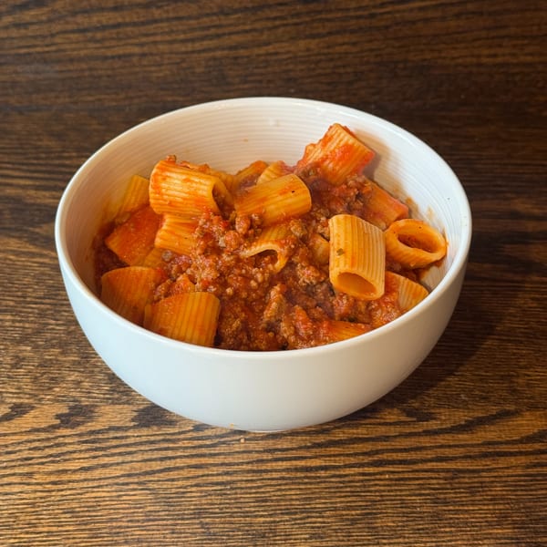 A white bowl filled with rigatoni pasta and a rich meat sauce, on a wooden table.
