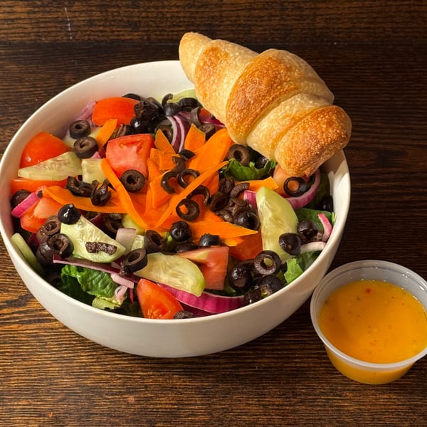 A fresh garden salad with tomatoes, cucumbers, olives, and carrots, served with a croissant and dressing.