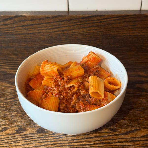 A white bowl filled with rigatoni pasta and a rich meat sauce on a wooden table.