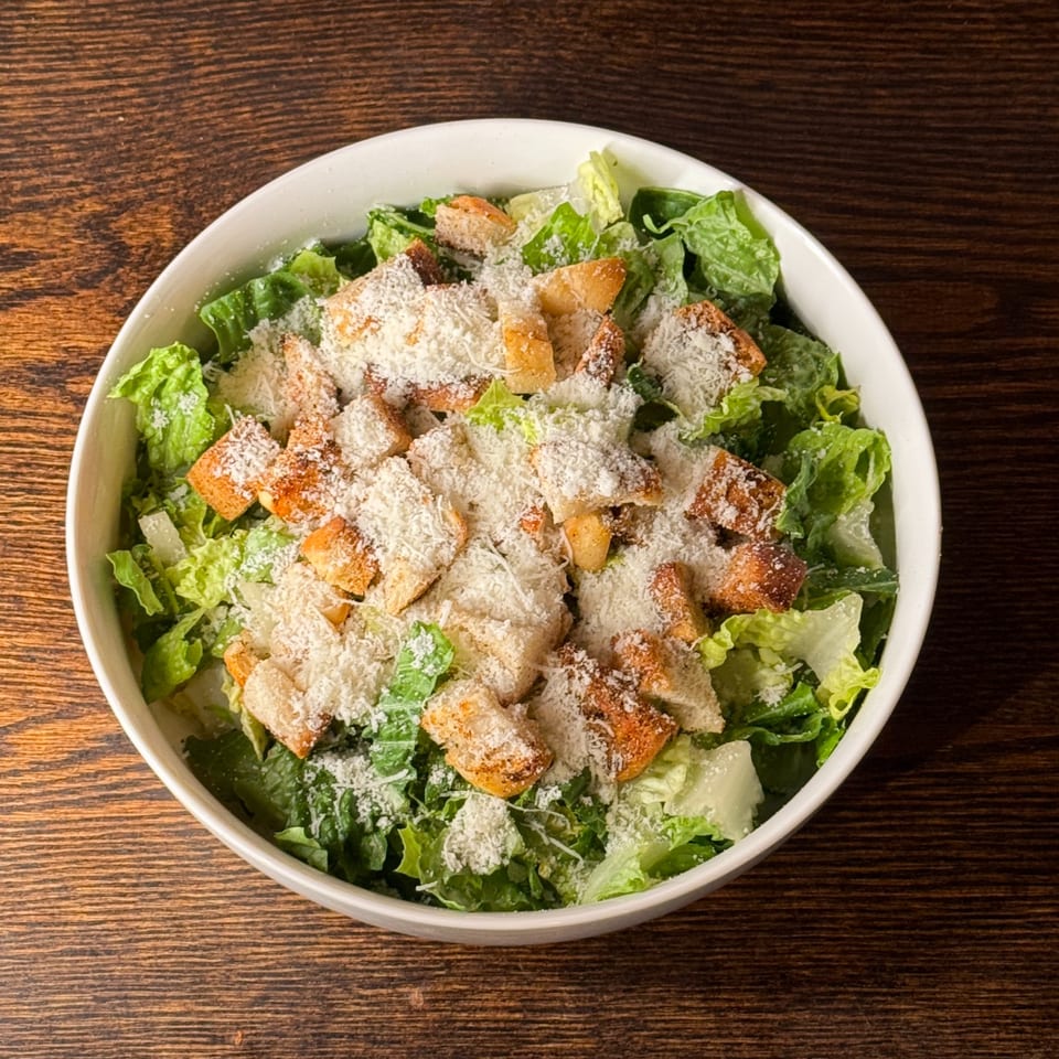 A white bowl of Caesar salad with romaine lettuce, croutons, and grated Parmesan cheese.