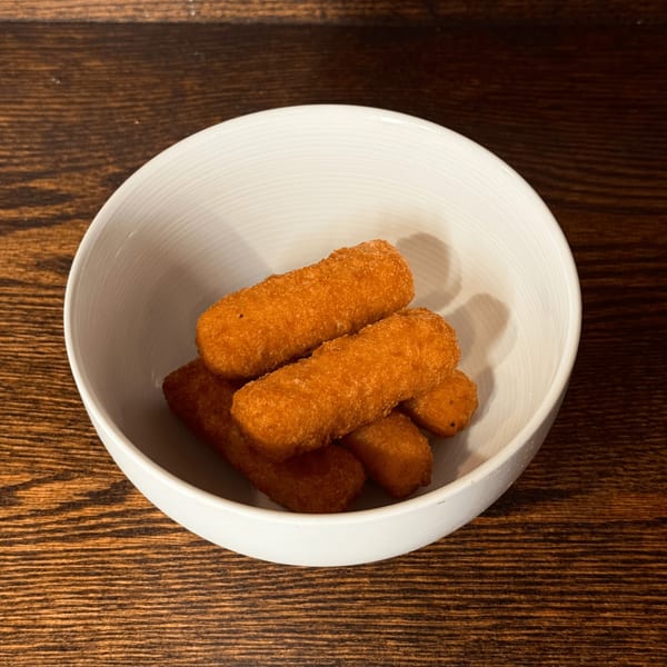 A white bowl filled with several golden-brown, crispy mozzarella sticks on a dark wooden table.