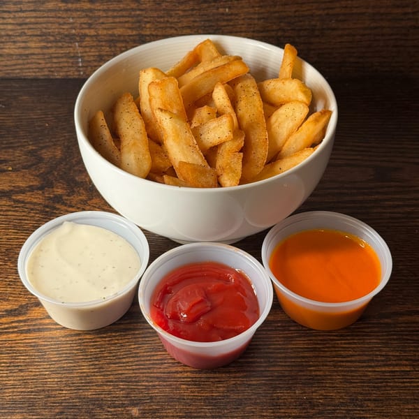 A white bowl of seasoned french fries with three dipping sauces on a wooden table.