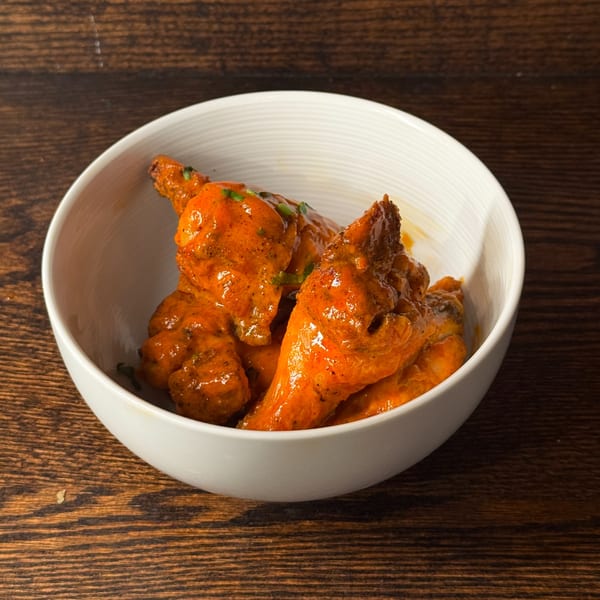 A white bowl filled with orange-sauced chicken wings, garnished with green herbs, on a wooden table.